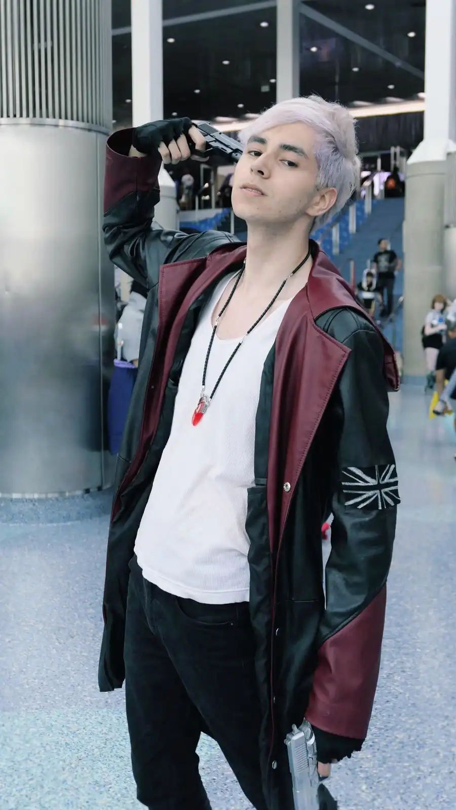 A person with light purple hair, wearing a black-and-maroon jacket with a Union Jack patch on the sleeve, a white t-shirt…