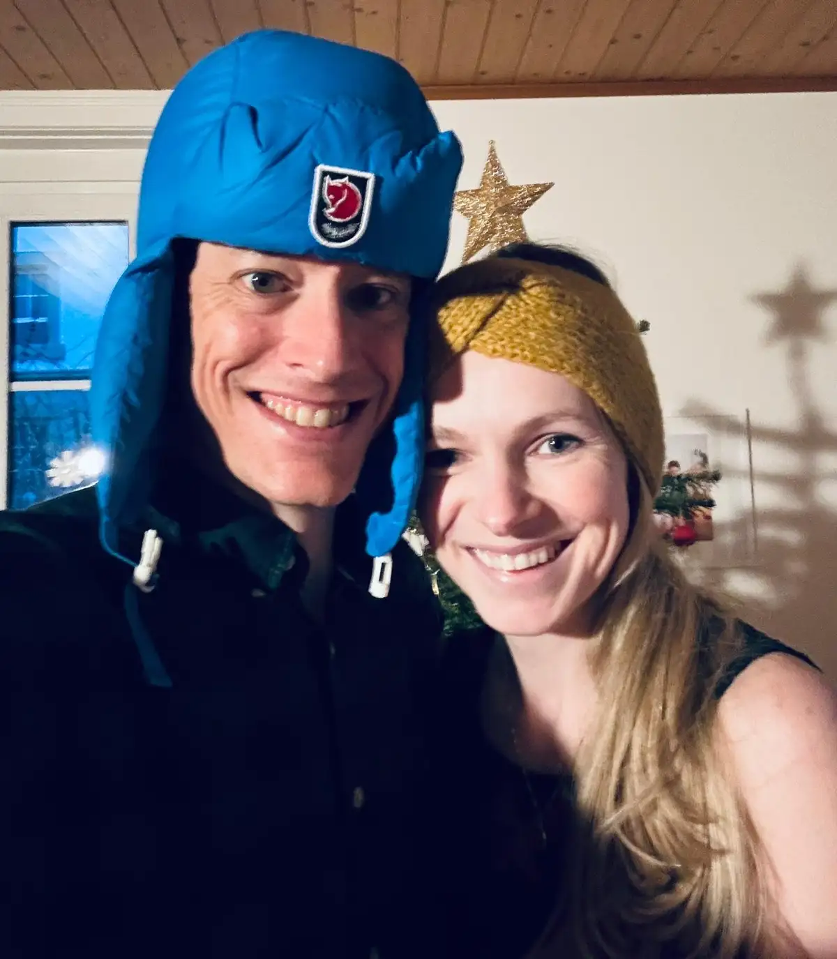 A smiling man and woman pose closely for a selfie indoors. The man wears a bright blue, puffy winter hat with ear flaps and…