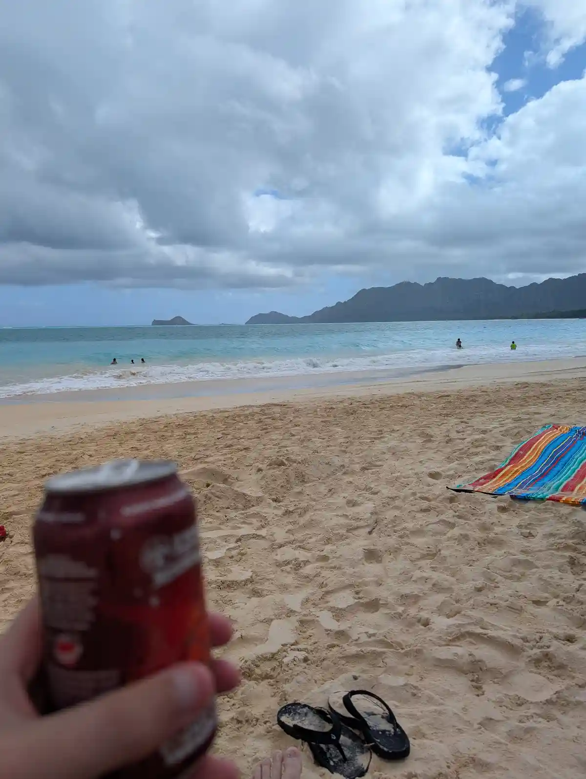 A first-person view from a beach with a hand holding a red soda can in the foreground, sandy ground with black flip-flops…