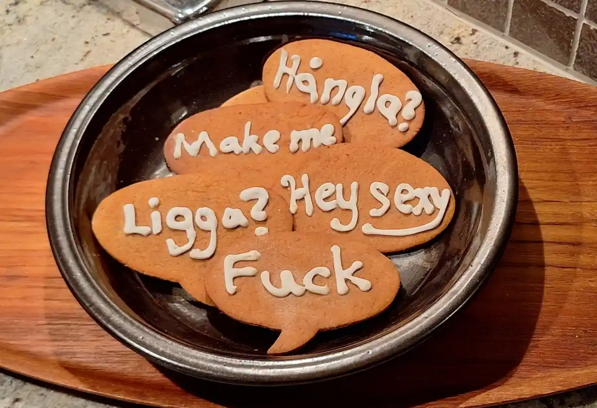Several gingerbread cookies shaped like speech bubbles sit in a dark ceramic bowl on a wooden board. Each cookie has white…