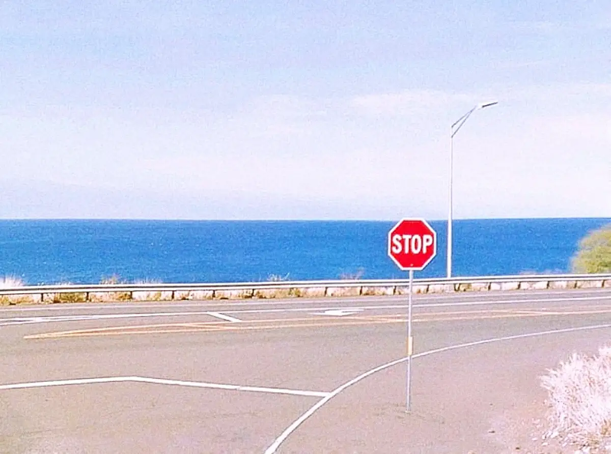 A red octagonal stop sign stands at a roadside intersection overlooking a vast blue ocean under a pale sky. The road has…
