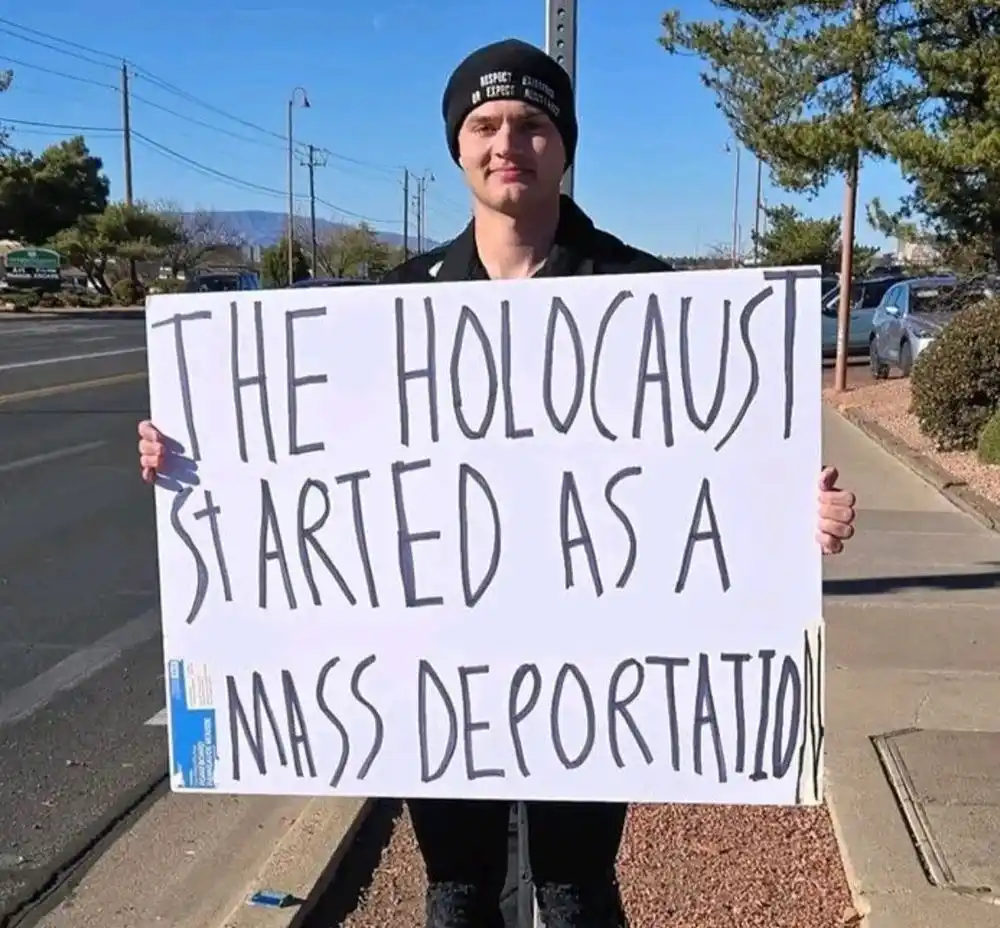 A young man wearing a black beanie stands outdoors on a sidewalk holding a large white sign with handwritten text. The sign…