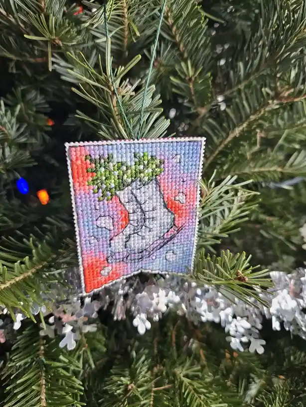 A close-up photo shows a handmade cross-stitch Christmas ornament hanging on a green pine tree. The ornament is…