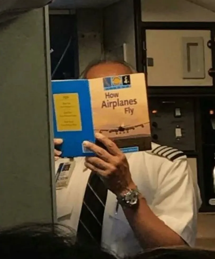 A pilot in a cockpit uniform holds up a book titled 'How Airplanes Fly' to cover his face, humorously implying he's not an…