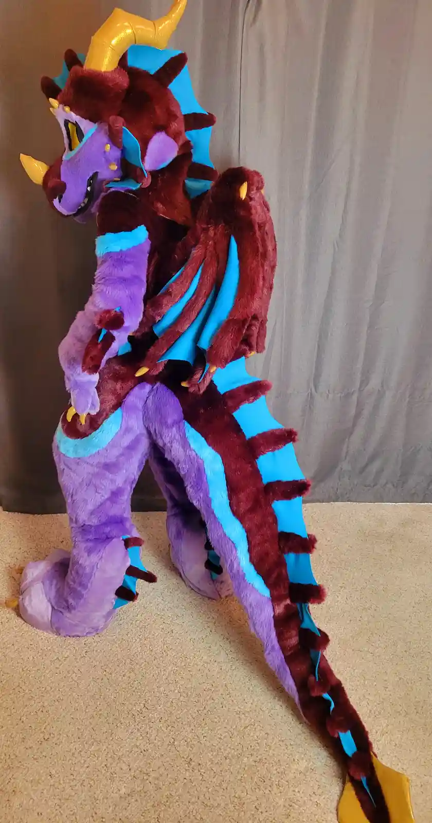 A person in a full-body, plush dragon costume stands on carpeted flooring, featuring purple and maroon fur, blue accents…