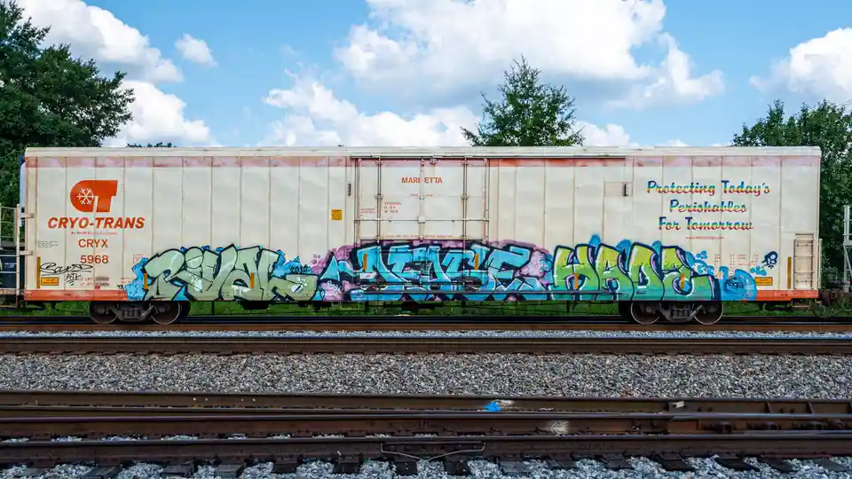 A side view of a white Cryo-Trans refrigerated railcar with colorful graffiti along its lower half, parked on tracks under a…