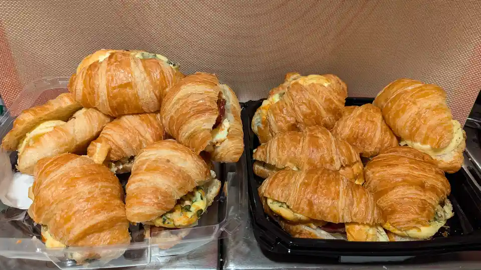 Two black and clear plastic containers are filled with golden-brown, flaky croissants filled with egg, cheese, and spinach…
