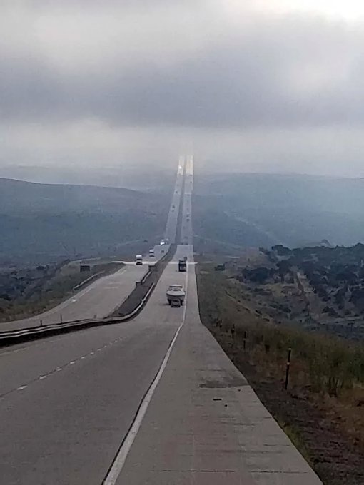 A long, straight highway stretches into a hazy, cloudy distance, flanked by hills and sparse vegetation. Several cars are…