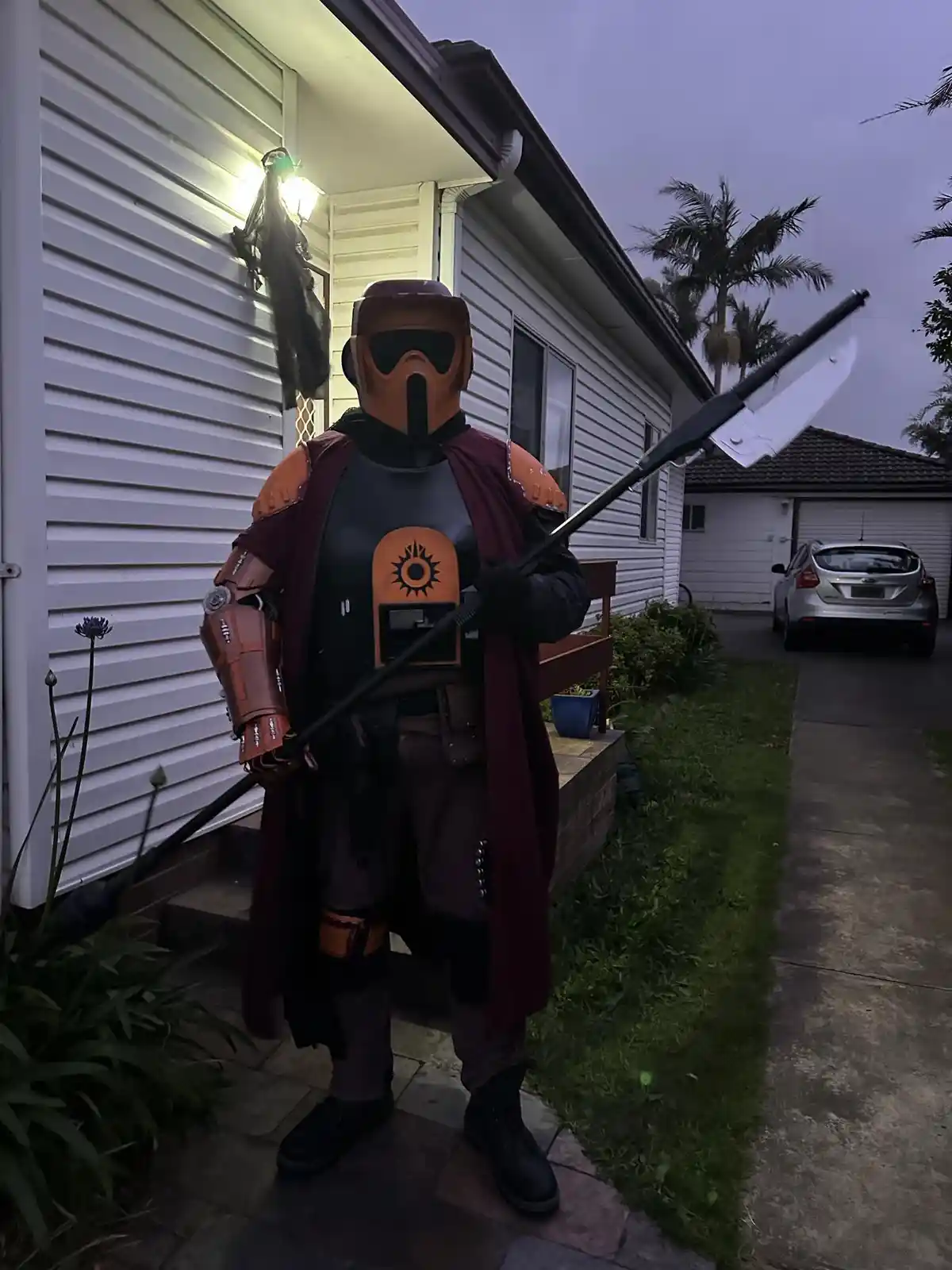 A person in a detailed Mandalorian-style costume, complete with helmet, cape, and a large polearm weapon, stands on a…