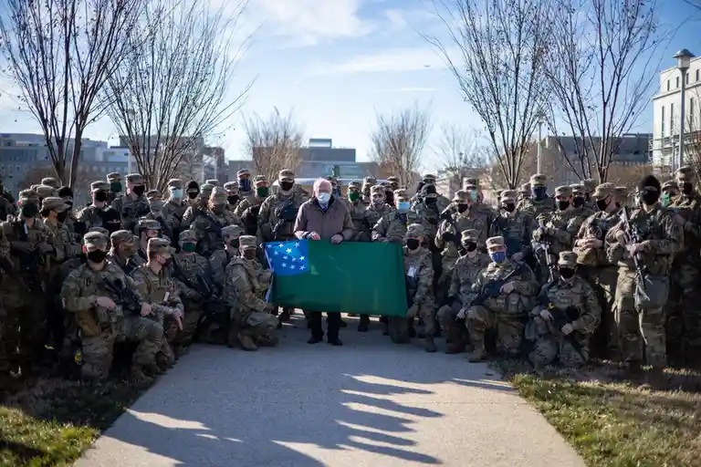 A man, identified as Bernie Sanders, stands in the center of a large group of U.S. soldiers in camouflage uniforms and face…