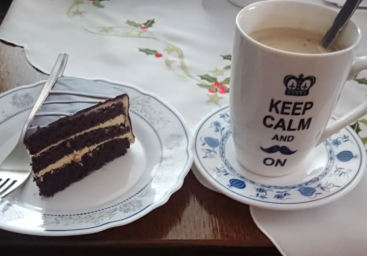 A slice of chocolate cake with cream layers sits on a blue-patterned plate next to a white mug with the text 'KEEP CALM AND…