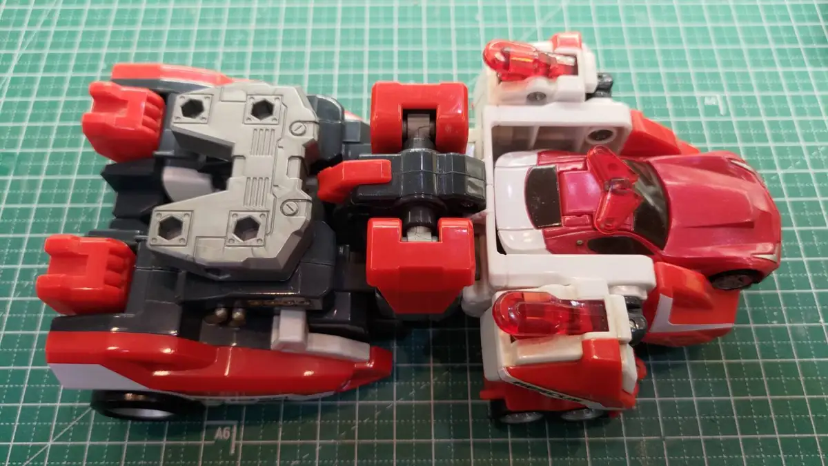 A red, white, and black toy robot, likely a transformer, is shown in its vehicle mode on a green cutting mat. The toy has…