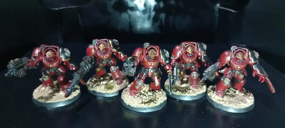 Five painted red and gold Space Marine miniatures stand in a row on textured bases, each holding different weapons. The…
