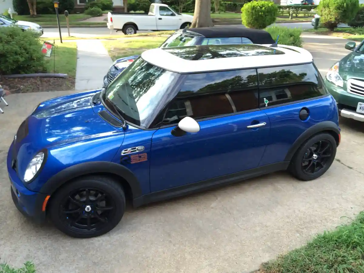 A blue Mini Cooper R53 with a white roof is parked in a driveway, featuring black wheels and trim. The car has a 'R53' badge…