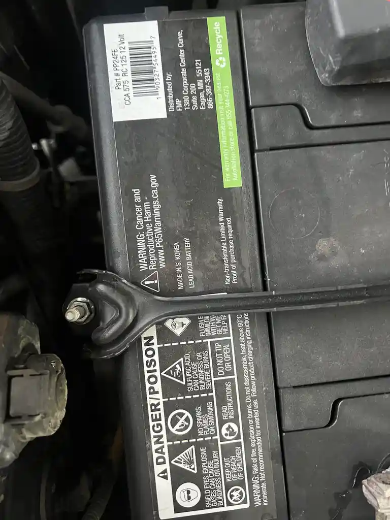 A close-up photo of a car battery with a large warning label showing 'DANGER/POISON' and various safety icons. A black…
