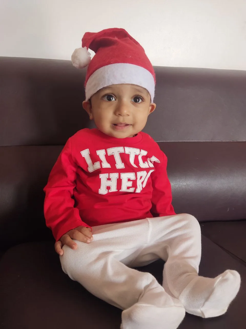 A baby wearing a red and white Santa hat and a red long-sleeved shirt with the words 'LITTLE HERO' in white block letters…
