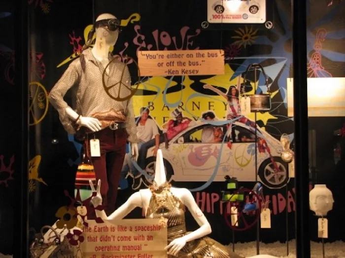 A retail window display features mannequins in 1960s hippie-style clothing, including peace signs and sunglasses, with a…