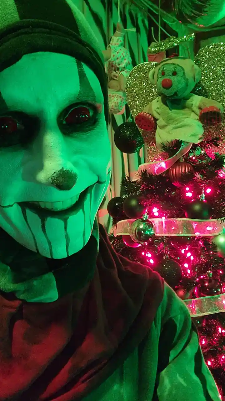A person with white face paint, black clown makeup, and red eyes poses beside a brightly lit pink Christmas tree. A…