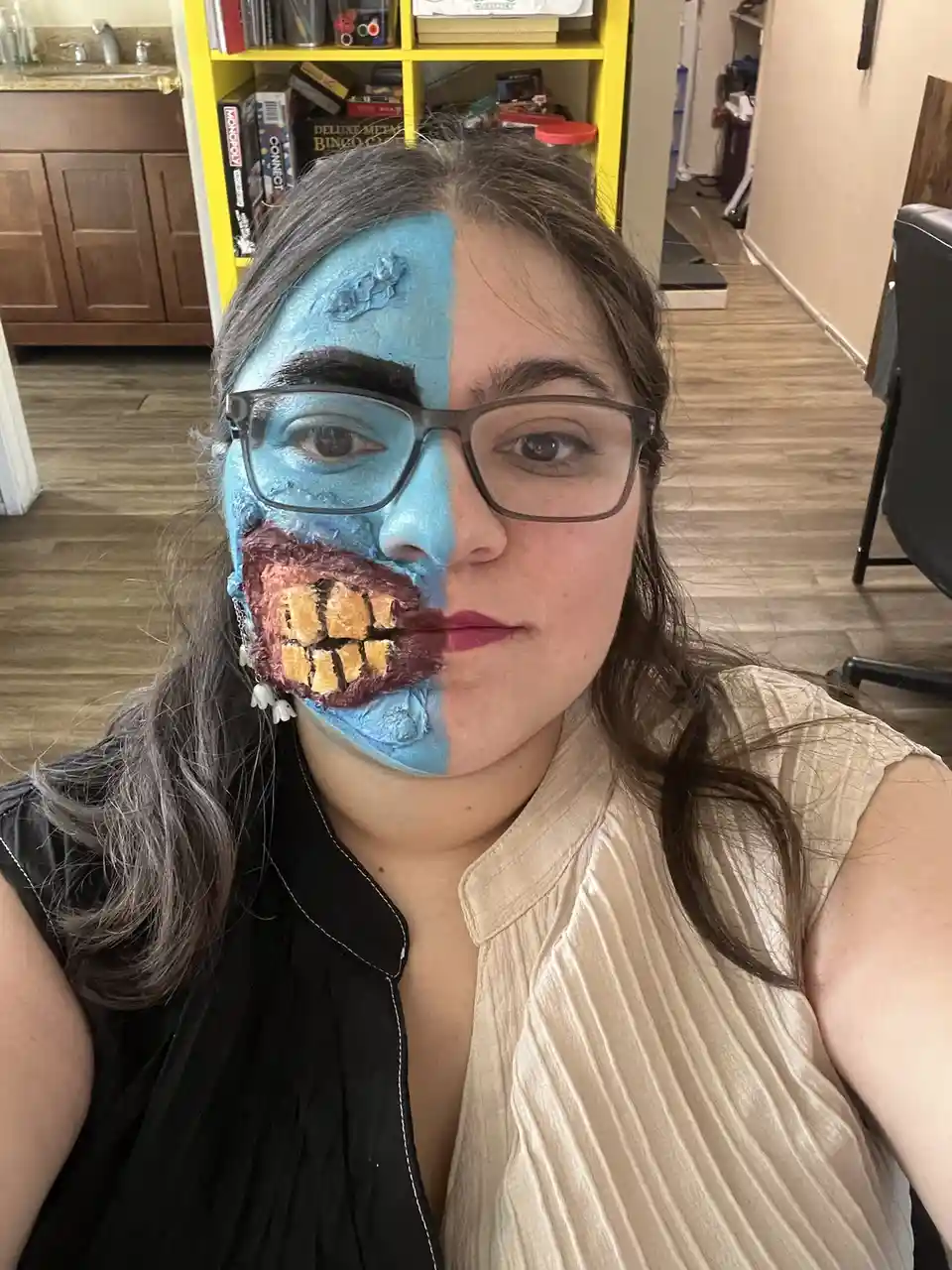 A person with glasses has their face painted half blue with a monster mouth and half natural skin, looking at the camera…