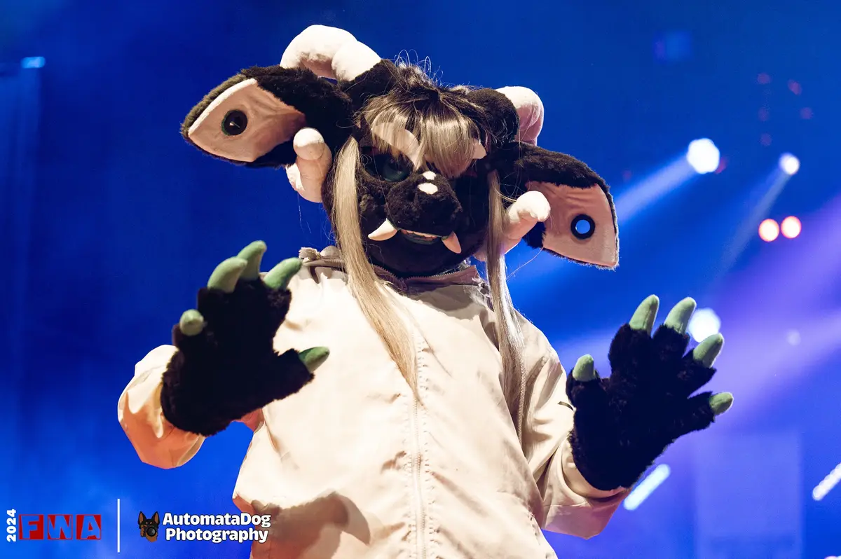 A person in a large, black, furry monster costume with oversized ears, fangs, and green-tipped claws, wearing a white jacket…