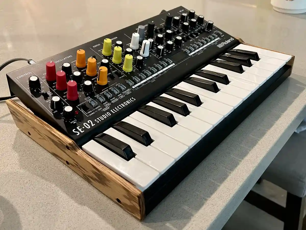 A Roland SE-02 Studio Electronics synthesizer with a wooden base sits on a light gray countertop, its black and white keys…