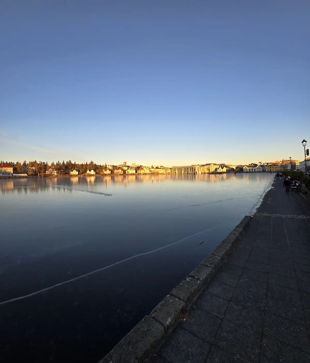 A wide shot of a frozen lake at sunset, with a city skyline reflected in the ice. A paved walkway runs along the right edge…