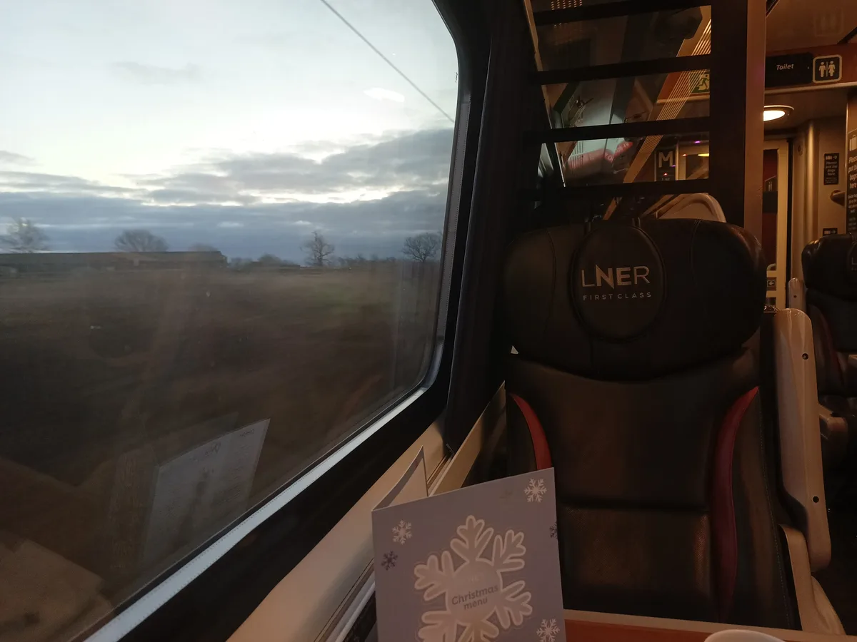 A photo taken from inside a moving train shows a black leather first-class seat with 'LNER FIRST CLASS' embroidered on the…