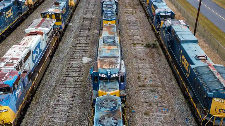 An aerial view shows multiple rusty CSX locomotives parked on parallel railroad tracks in a yard, with one central engine…