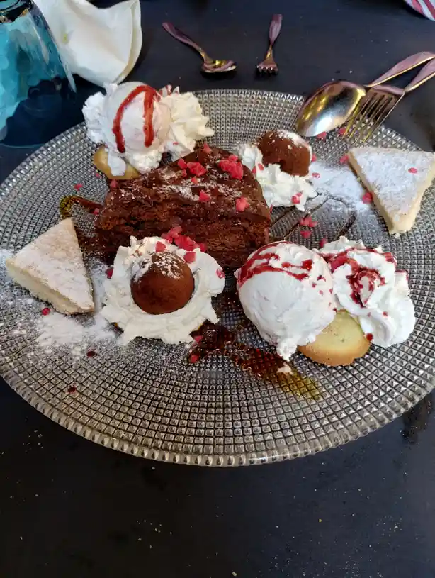 A plate overflowing with various desserts including chocolate cake, ice cream scoops, whipped cream, powdered sugar, and…