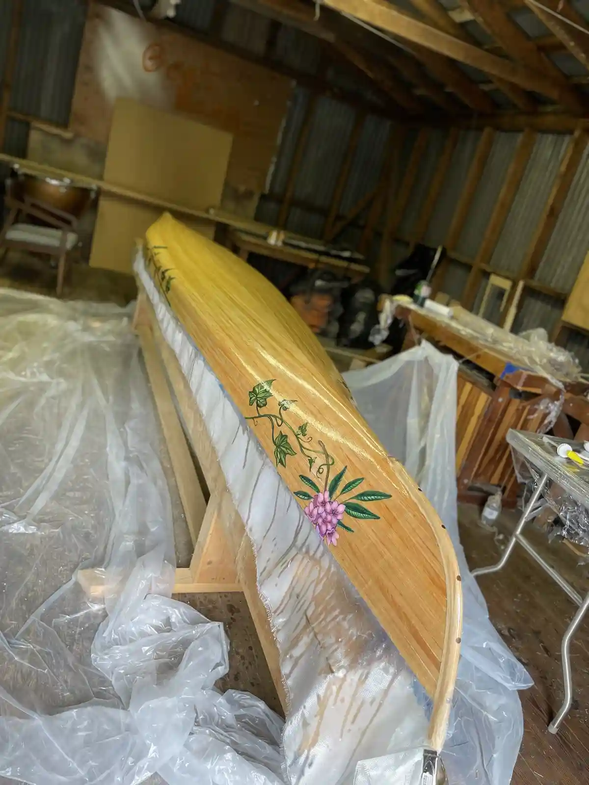 A wooden canoe rests on a stand inside a workshop, its side decorated with a painted floral design featuring pink flowers…
