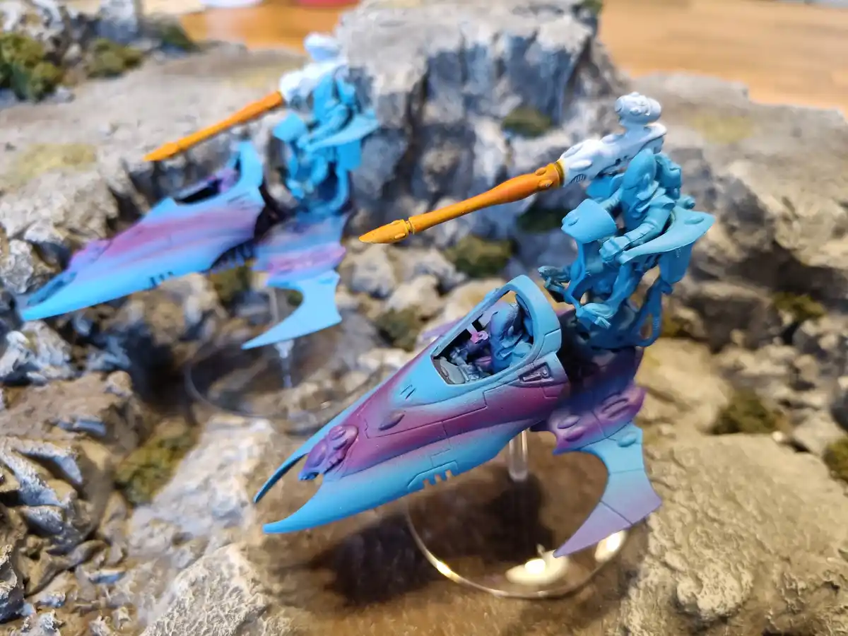 Two painted miniature sci-fi fighter craft with blue and purple gradient finishes sit on rocky terrain. Each has a pilot and…