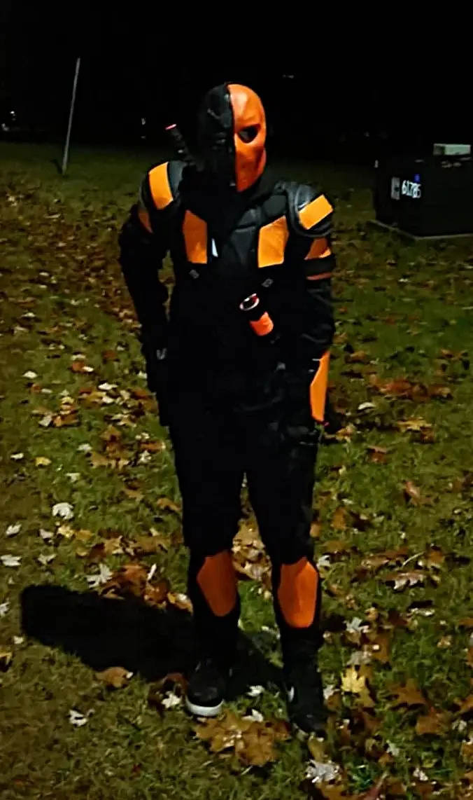 A person in a full-body costume resembling the comic book character Deathstroke stands outdoors at night on grass covered…