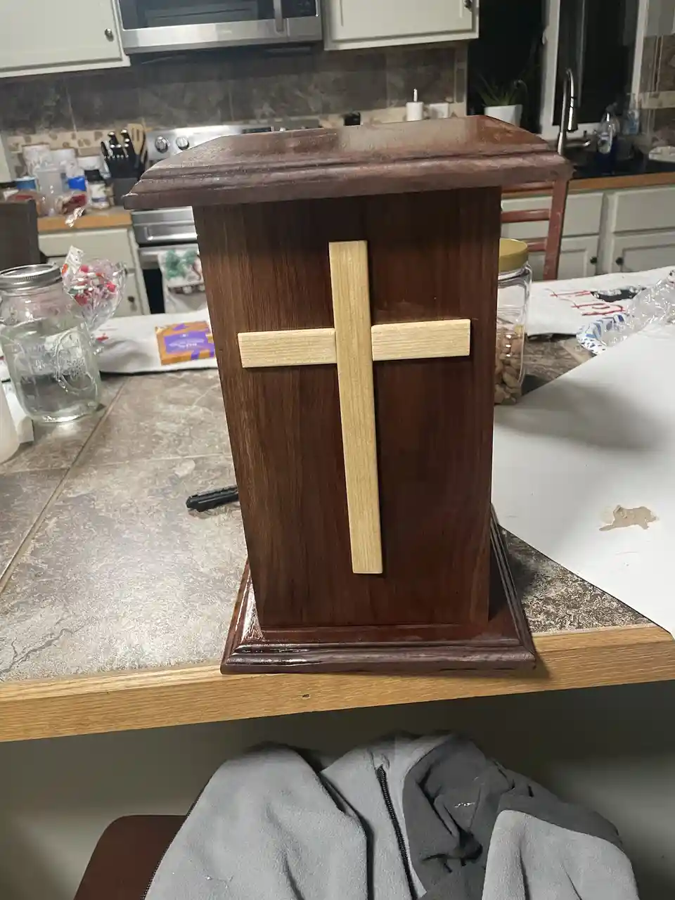 A dark brown wooden box with a light-colored wooden cross affixed to its front sits on a kitchen counter. The background…