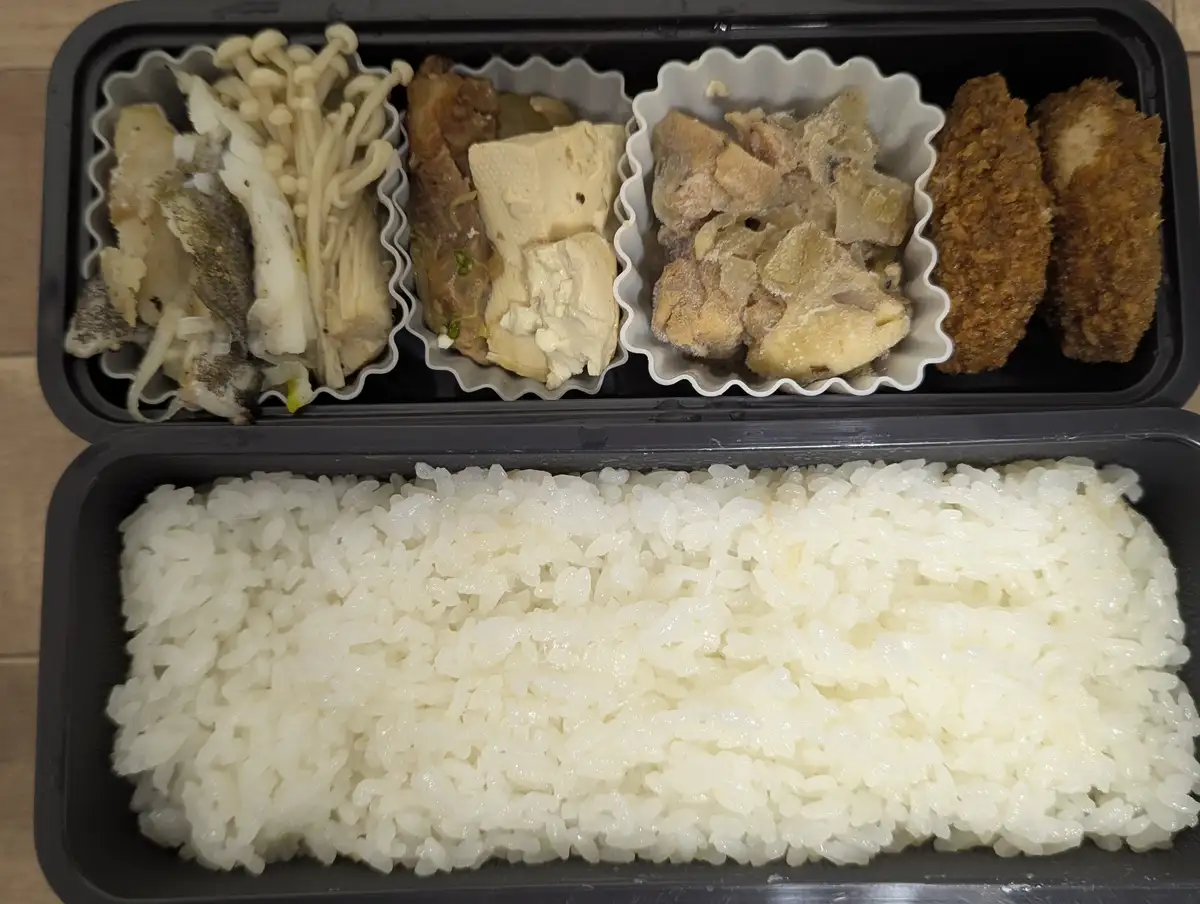 A top-down photo of a black bento box with a large compartment of white rice at the bottom and four smaller compartments…