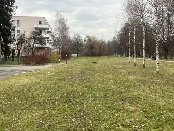 A wide shot of a grassy park with bare trees, a path, and apartment buildings in the background under an overcast sky. The…