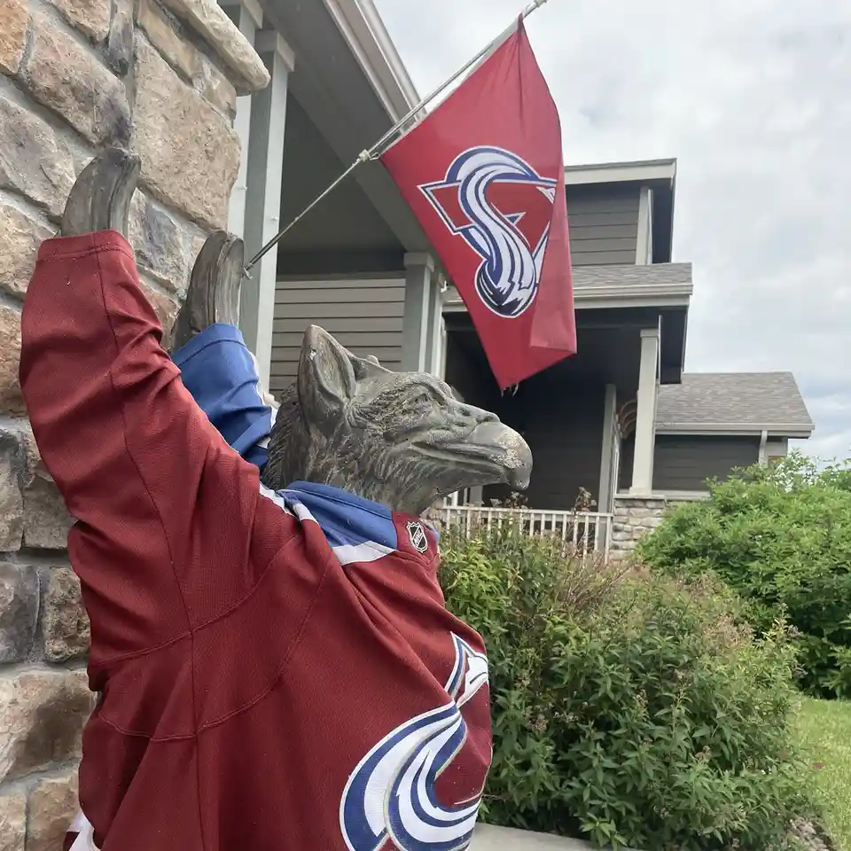 A stone gargoyle statue wearing a Colorado Avalanche NHL hockey jersey with arms raised in celebration, positioned beside a…