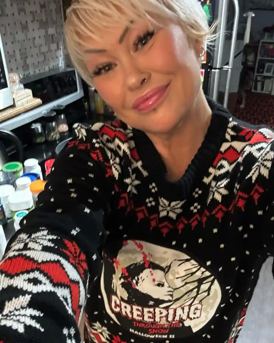 A person with short blonde hair and makeup takes a selfie in a kitchen, wearing a black, red, and white patterned sweater…