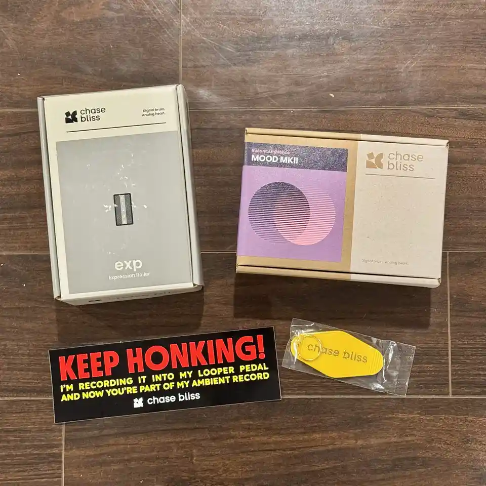 Two Chase Bliss audio effect pedal boxes, 'exp Expression Roller' and 'Mood MKII', sit on a wooden floor next to a yellow…