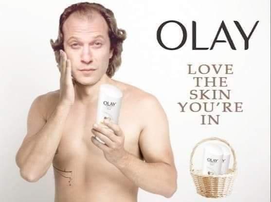A shirtless man with curly hair holds an Olay cream tube while touching his cheek, beside text reading 'OLAY LOVE THE SKIN…