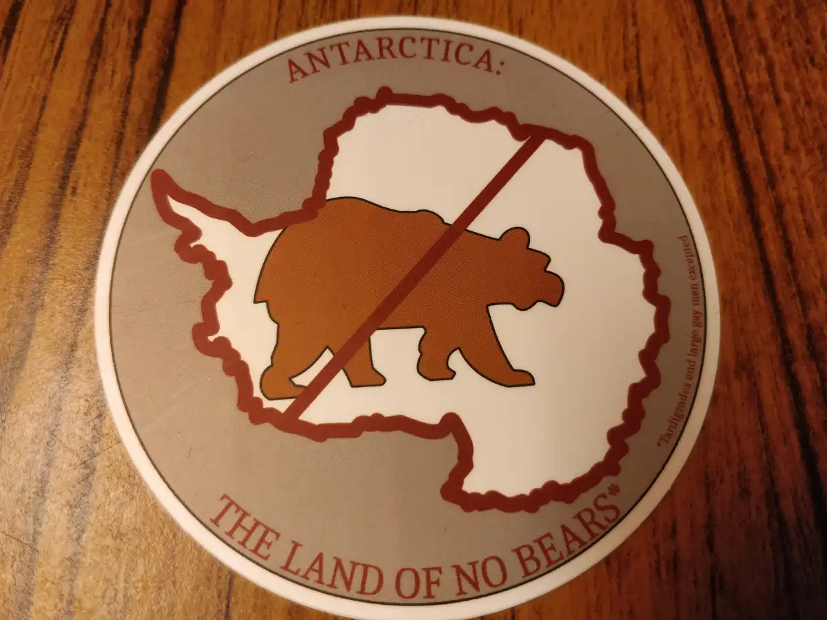 A circular sticker on a wooden surface shows a map outline of Antarctica with a brown bear silhouette crossed out by a red…