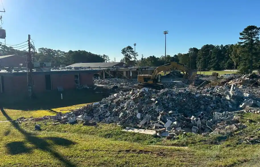 A yellow excavator is demolishing a brick building, leaving a large pile of rubble on a grassy field under a clear blue sky…