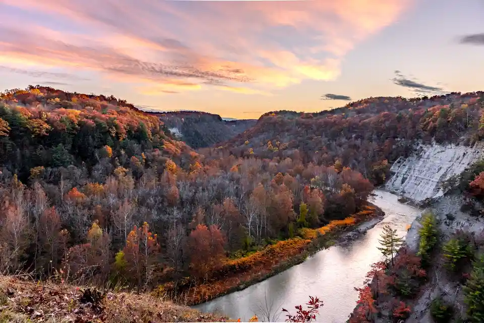 A scenic autumn landscape featuring a river winding through a valley flanked by hills covered in colorful foliage under a…