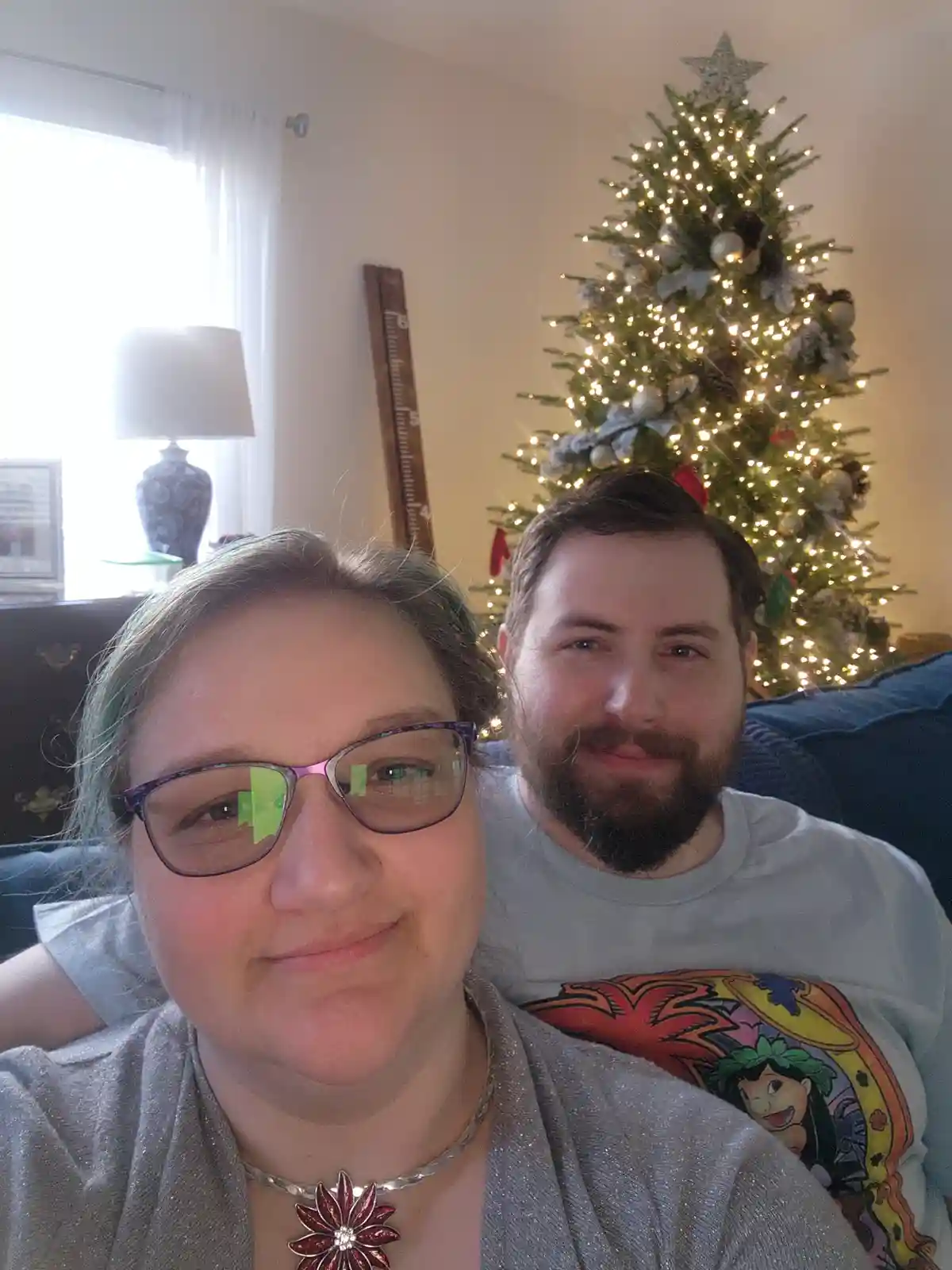 A selfie of a woman with glasses and a man with a beard sitting on a couch in front of a lit Christmas tree. The woman is in…