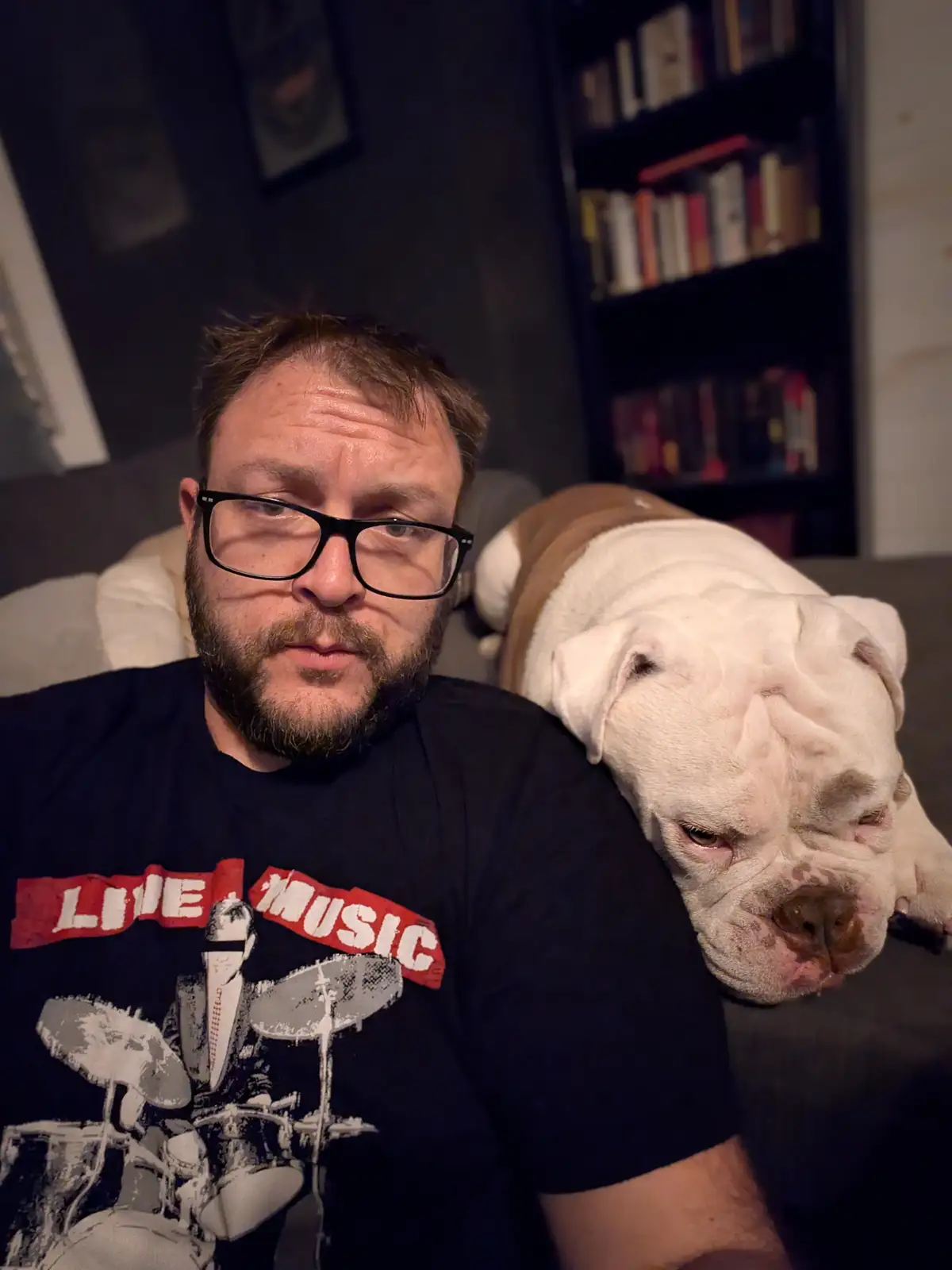 A man with glasses and a beard wearing a 'LIVE MUSIC' t-shirt sits on a couch with a large white and brown bulldog resting…