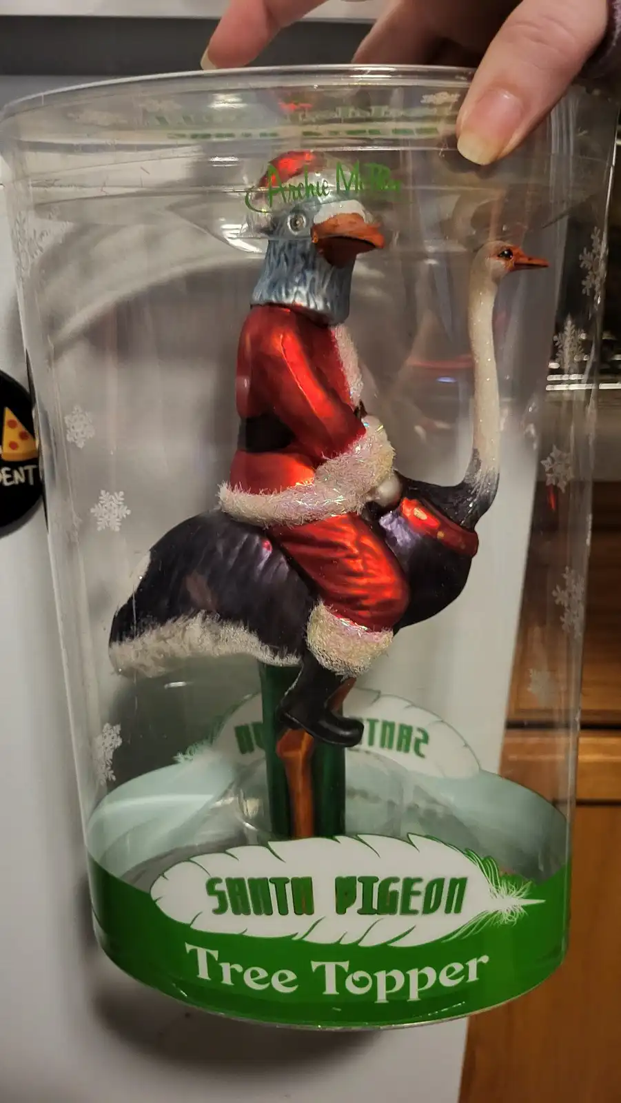 A plastic Christmas tree topper ornament in its packaging shows a bird with a blue head and orange beak dressed in a red…