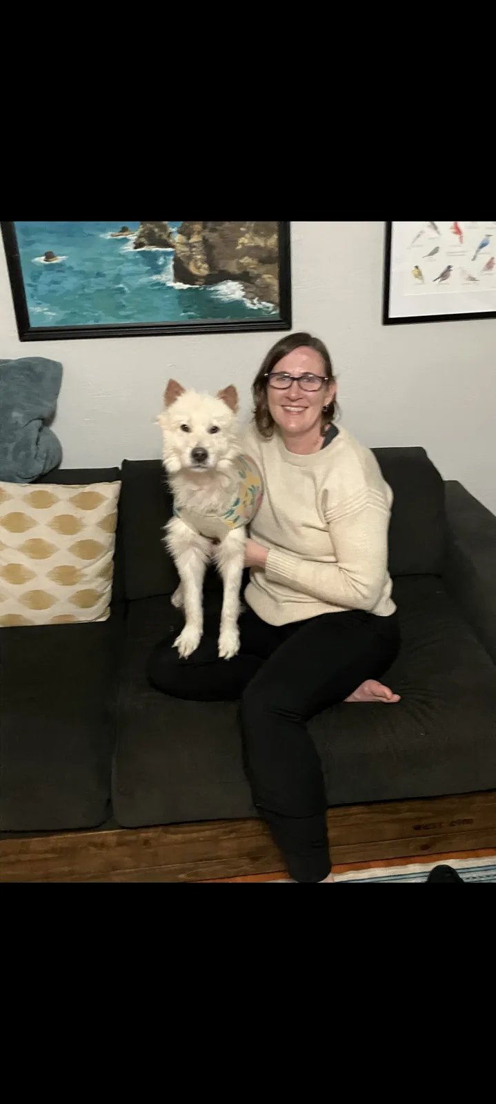 A woman with glasses and a cream sweater sits on a dark couch holding a fluffy white dog wearing a patterned vest. The dog…