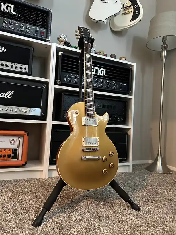 A gold-finished electric guitar stands upright on a black tripod stand in a room with carpeted flooring. Behind it, a white…
