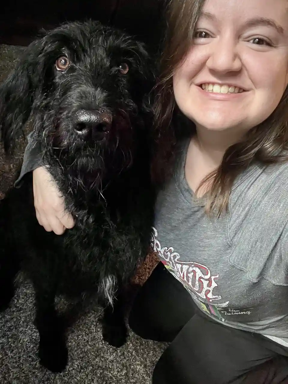A person with brown hair and a grey t-shirt is smiling while holding a large black shaggy dog that looks directly at the…