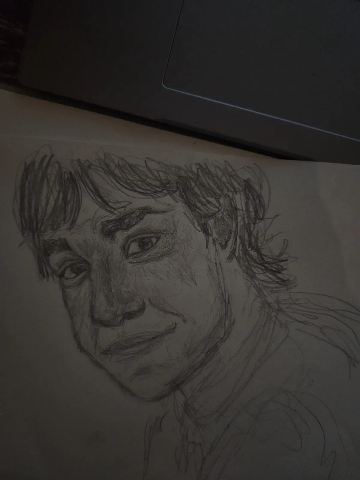 A dimly lit pencil sketch of a person's face with messy hair and a neutral expression, partially obscured by a laptop lid…