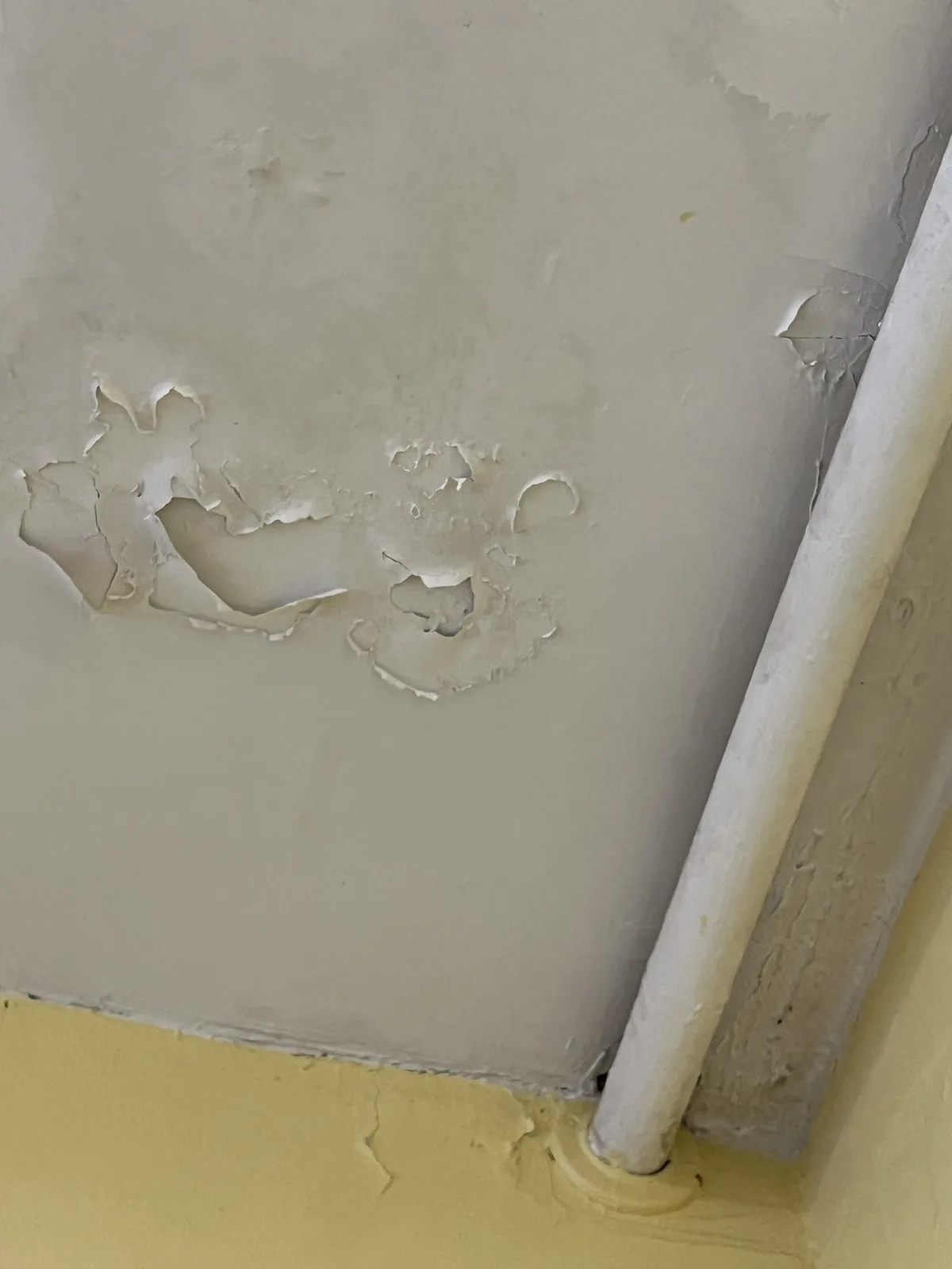 A close-up photo shows a wall with peeling, damaged beige paint revealing the underlying surface, next to a white pipe and a…